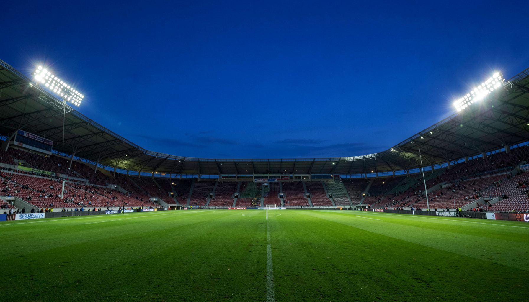 A wide stadium at dusk with floodlights glowing against a deep blue sky, green pitch stretching across the frame