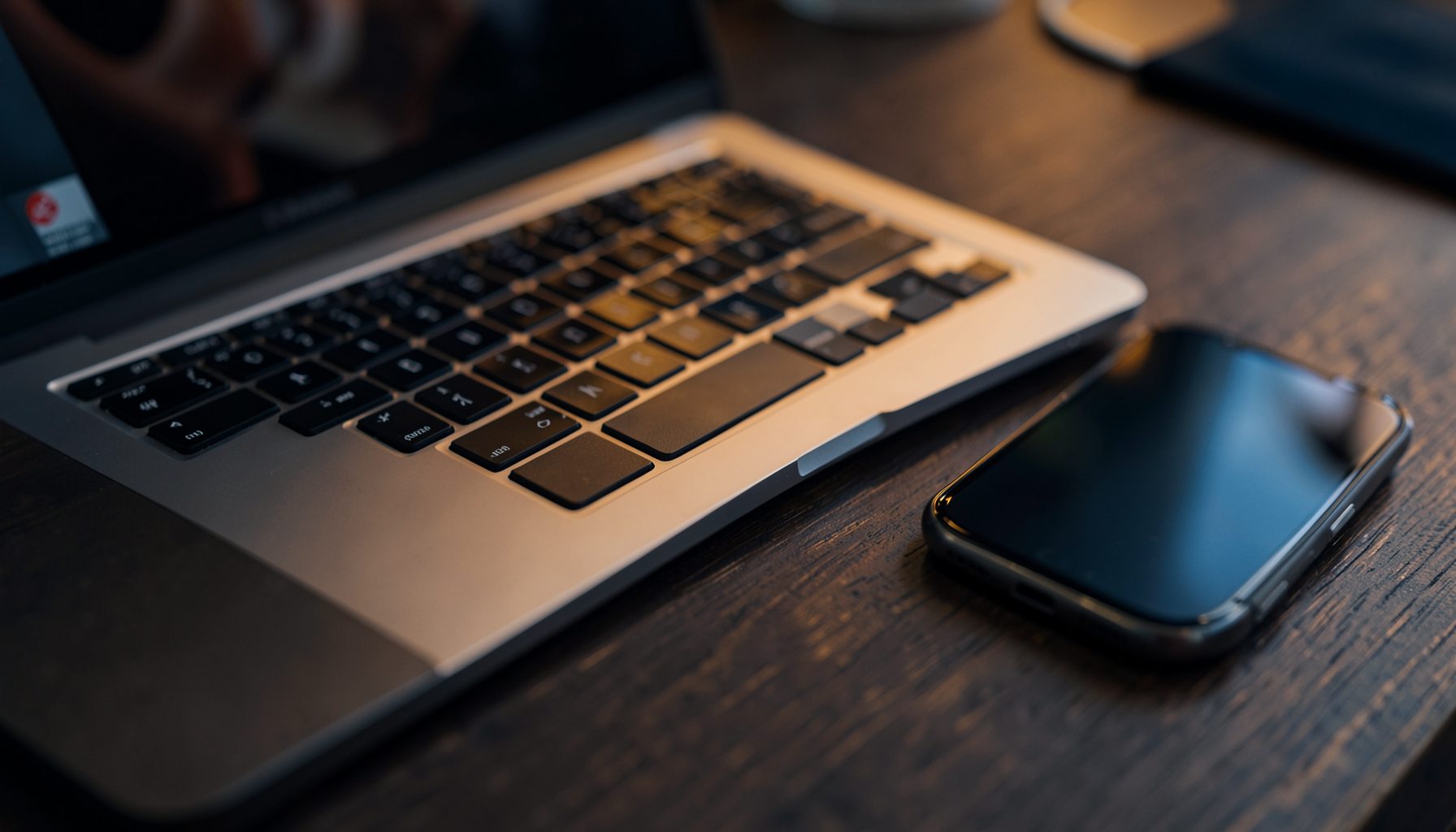 Close-up of a sleek laptop keyboard with a smartphone beside it on a dark wooden desk, warm amber and navy tones